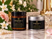 Load image into Gallery viewer, Two scented candles from the Powder &amp; Pearls Cherish Collection are displayed on a marble surface with decorative flowers, pearls, and gold-toned background elements. One candle is in an amber glass jar with a black lid, and the other is in a silver tin with a black lid.